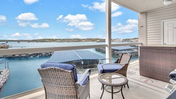 Een balkon met rieten meubilair, een glazen tafel en uitzicht op een jachthaven en een waterpartij.