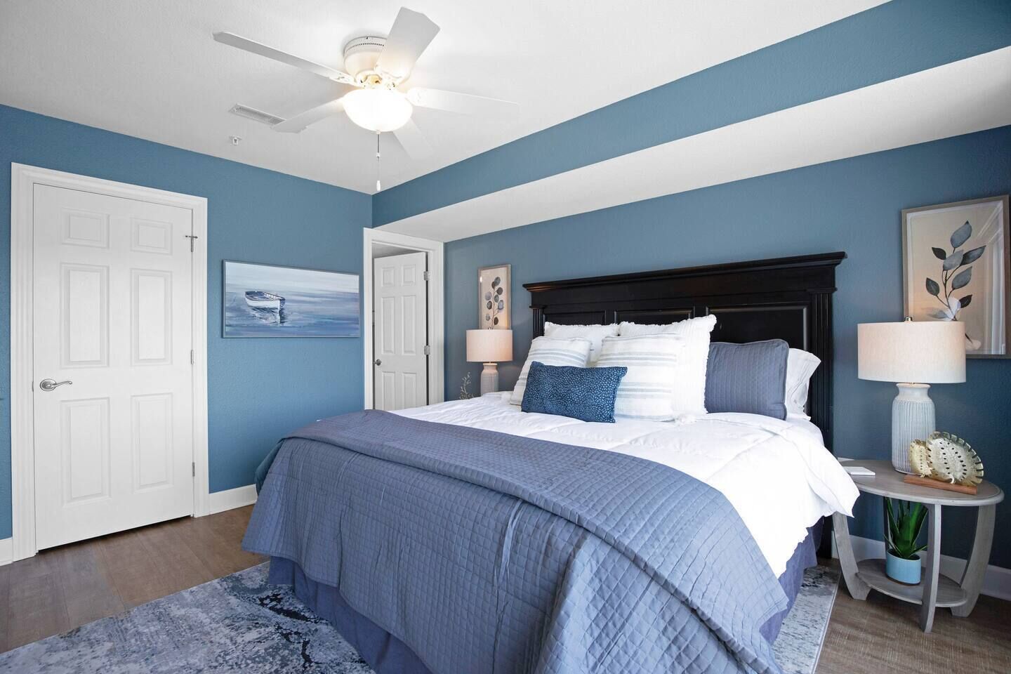 Ein Schlafzimmer mit einem Bett, einem Deckenventilator, einem Nachttisch, einer Lampe und einem gerahmten Bild an der Wand.