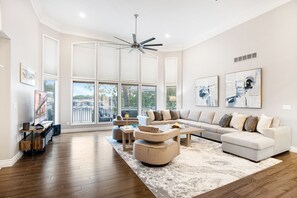 Un salon spacieux avec un grand canapé d’angle, une table basse et un ventilateur de plafond. De grandes fenêtres laissent entrer la lumière naturelle et offrent une vue sur l’extérieur.
