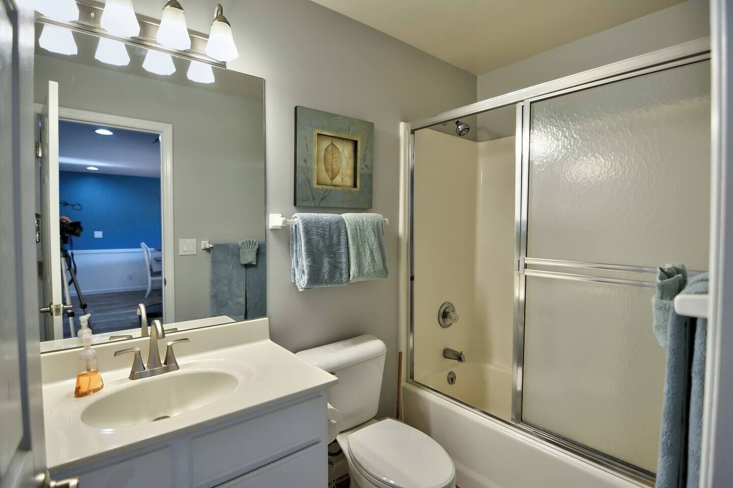 Une salle de bain avec un lavabo blanc, un miroir et des toilettes, une douche avec une porte en verre, et une serviette bleue accrochée au mur.