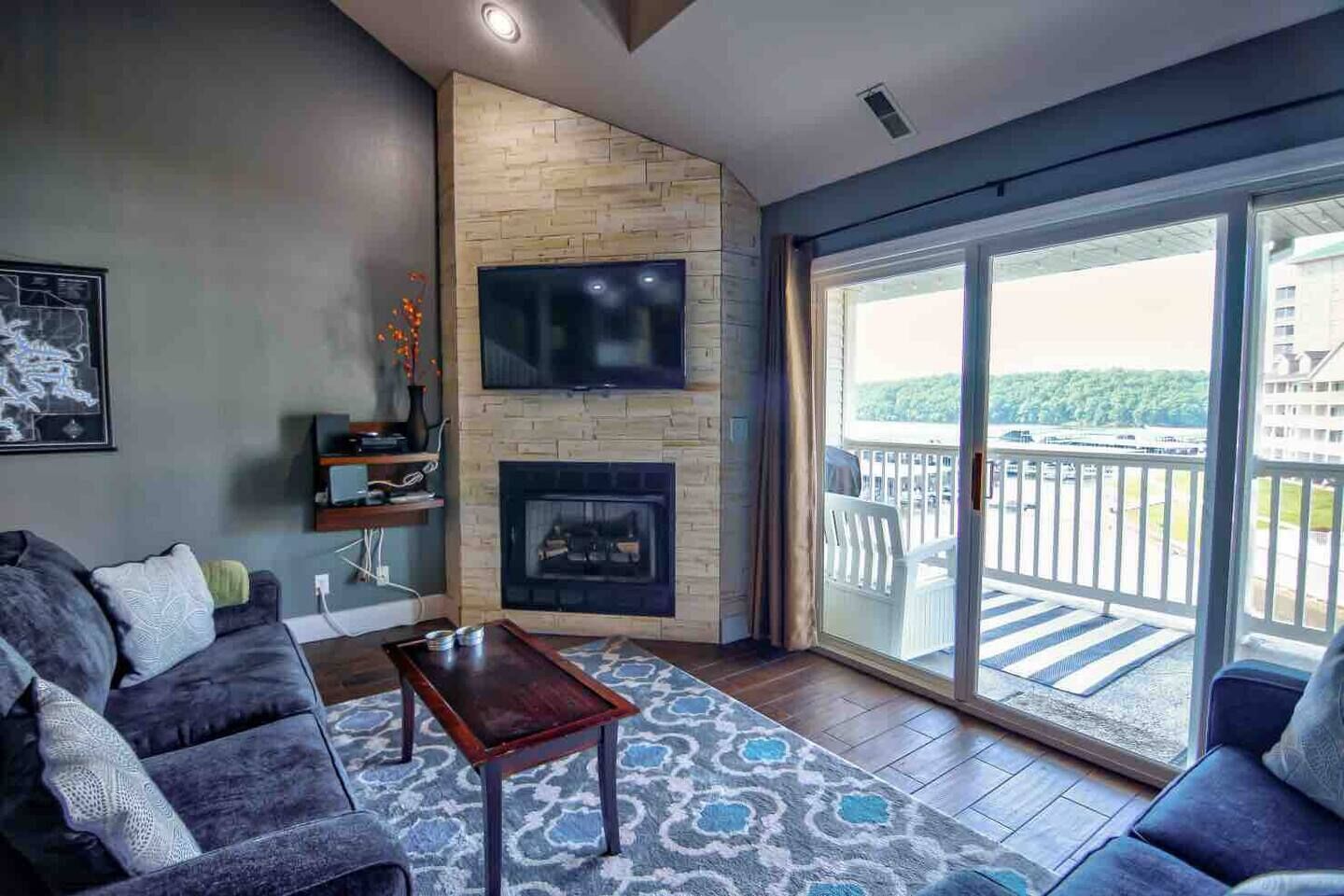 Una sala de estar moderna con chimenea, televisor de pantalla plana y balcón con vistas a un edificio y vegetación.