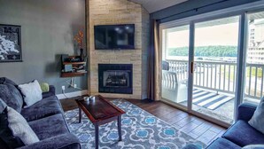 A modern living room with a fireplace, a flat-screen TV, and a balcony with a view of a building and greenery.