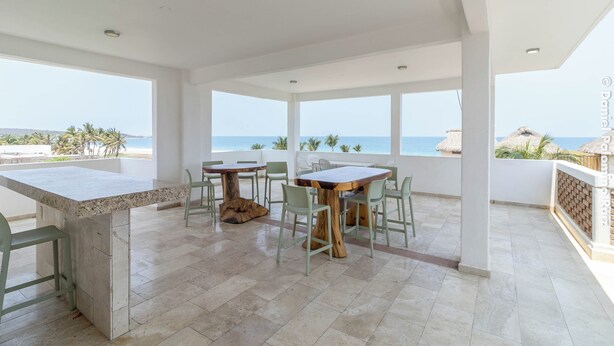 Ein geräumiger Balkon mit Essbereich, ausgestattet mit einem großen Tisch und Stühlen, mit Blick auf einen Strand mit Palmen und einer strohgedeckten Konstruktion.