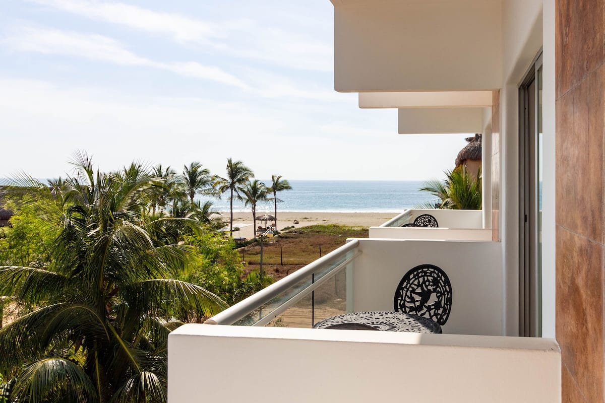 Un balcón con vista a la playa, palmeras y una construcción con techo de paja.