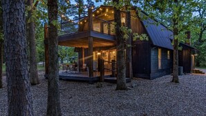 A cabin with a wooden deck, a covered outdoor seating area, and a large window overlooking the forest.