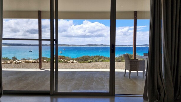 A room with a large sliding glass door leading to a balcony with a view of the sea and boats.
