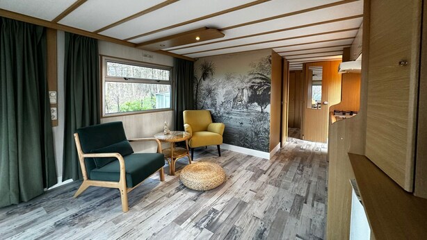 Ett rum med ett träpanelat tak, ett stort fönster med gardiner, en grön stol, en gul stol, ett litet runt bord och en vävd pall.