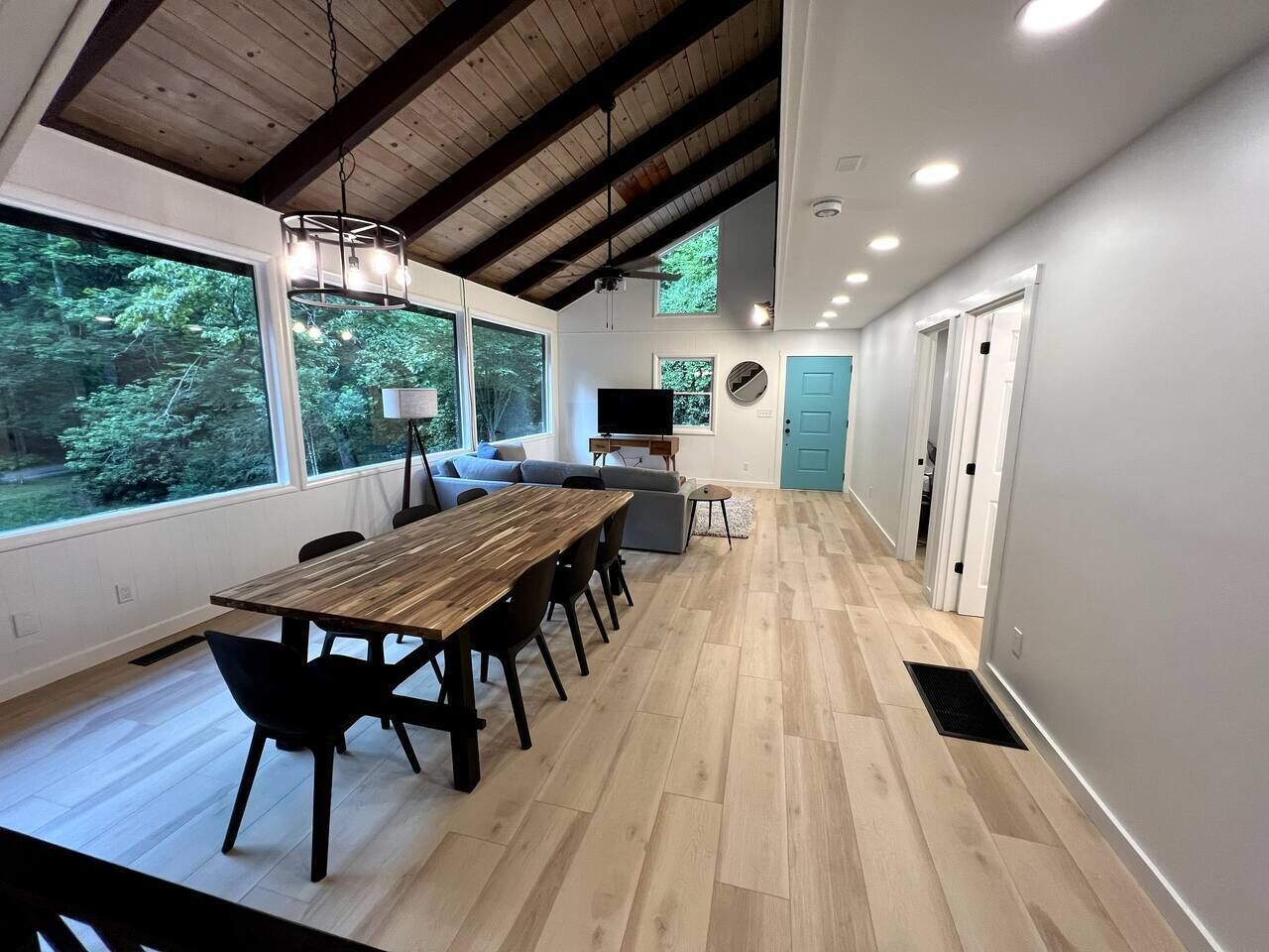 A spacious room with a wooden dining table, black chairs, a sofa, and a TV. Large windows offer a view of greenery outside.