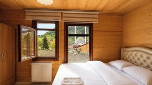 A bedroom with wooden walls, a bed with white bedding, a radiator, and two windows offering views of a landscape and a building. - Uzungol Cerrahpasa Suites (Çaykara)