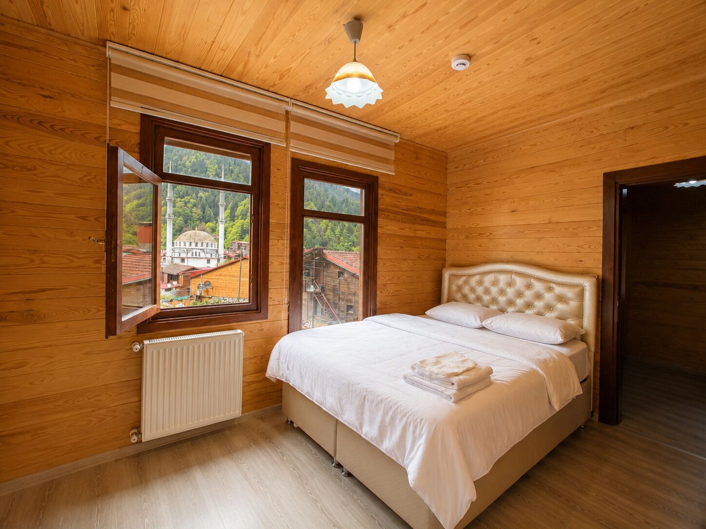 A bedroom with wooden walls, a bed with white bedding, and two windows offering a view of buildings and trees.