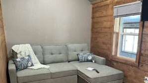 Una habitación de esquina con un sofá seccional gris y reposapiés, piso de madera, una ventana con persianas y una pared de madera.