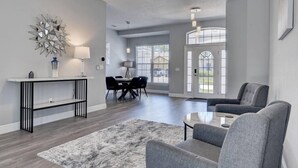 A modern living room with a fireplace, a dining area, and large windows. - Remodeled Golf Course Pool & Spa Home (Davenport)