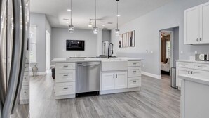 A modern kitchen with white cabinetry, stainless steel appliances, pendant lighting, and a flat-screen TV mounted on the wall. - Remodeled Golf Course Pool & Spa Home (Davenport)