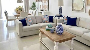Ein modernes Wohnzimmer mit einem beige farbenen Sofa, einem Holztisch und dekorativen Kissen. Im Hintergrund ist ein Essbereich mit Tisch und Stühlen zu sehen.