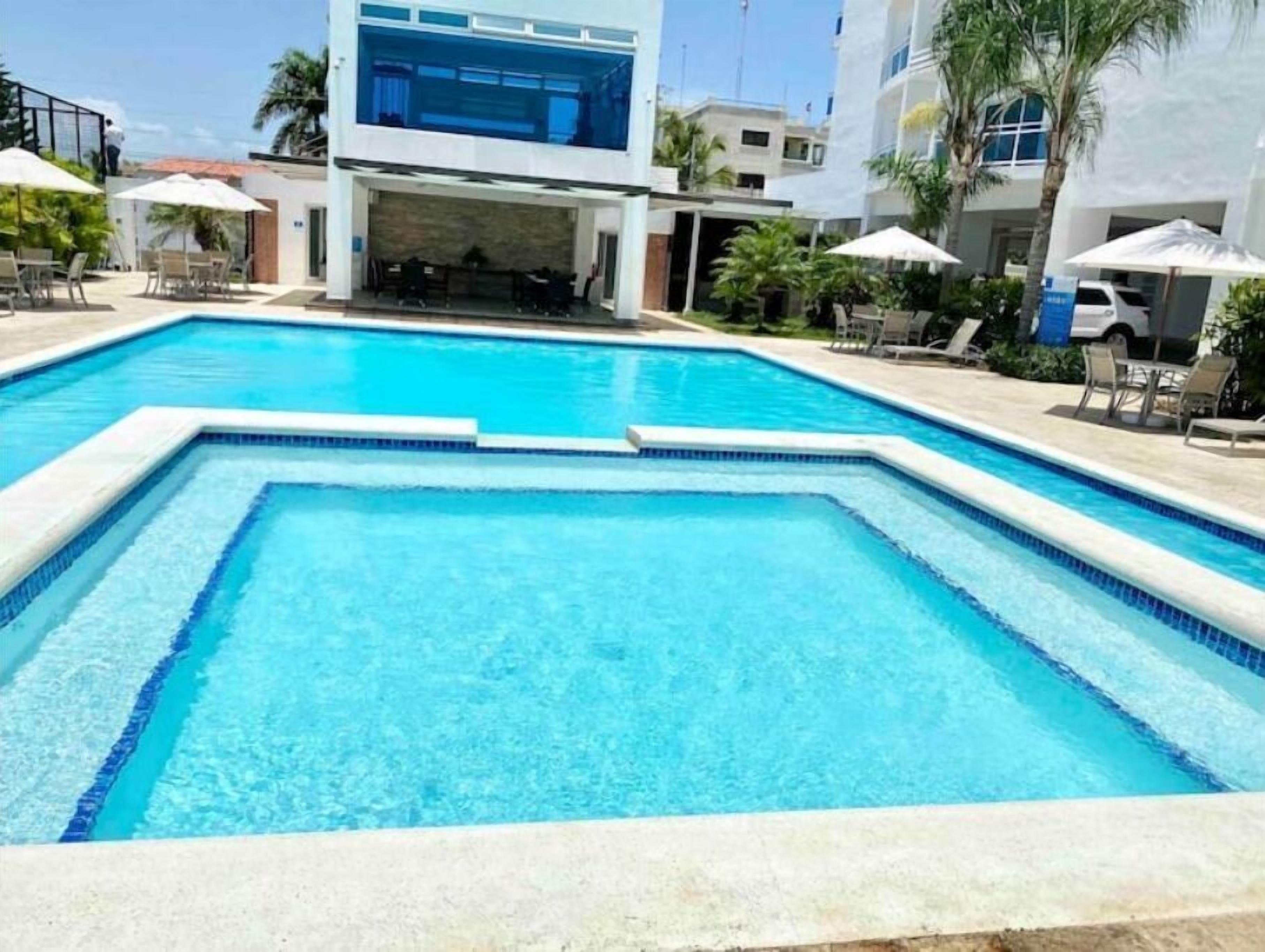 Een rechthoekig zwembad met helderblauw water, omringd door ligbedden en parasols, en grenzend aan moderne gebouwen met balkons.