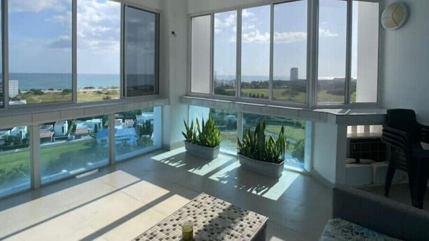 Amplia habitación con ventanas grandes que ofrecen vistas al mar y edificios. Hay plantas en macetas y un ventilador de techo.