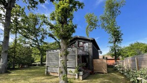 Une petite cabane en bois, dotée d’un toit en pente, entourée d’une cour verdoyante et d’arbres.