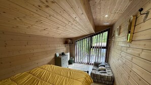 Une pièce de cabane en bois comprenant un lit, un banc avec des coussins, une petite table et une fenêtre avec des stores verticaux.