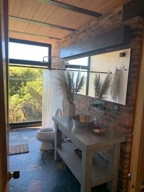 A bathroom with a modern design, featuring a large mirror, a sink, and a toilet. The room has a stone floor and a view of greenery outside.