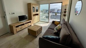 A modern living room with a flat-screen TV, wooden furniture, a sofa, and a large window offering a city view. - Quiberon - 2 rooms apartment - 40m² - quiet area (QUIBERON)