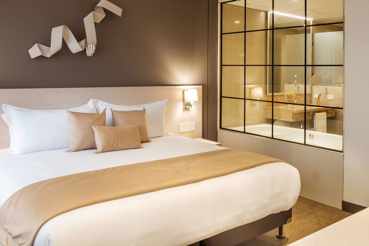 A hotel room with a large bed, a nightstand with a lamp, a glass-enclosed bathroom, and a wall-mounted abstract art piece.