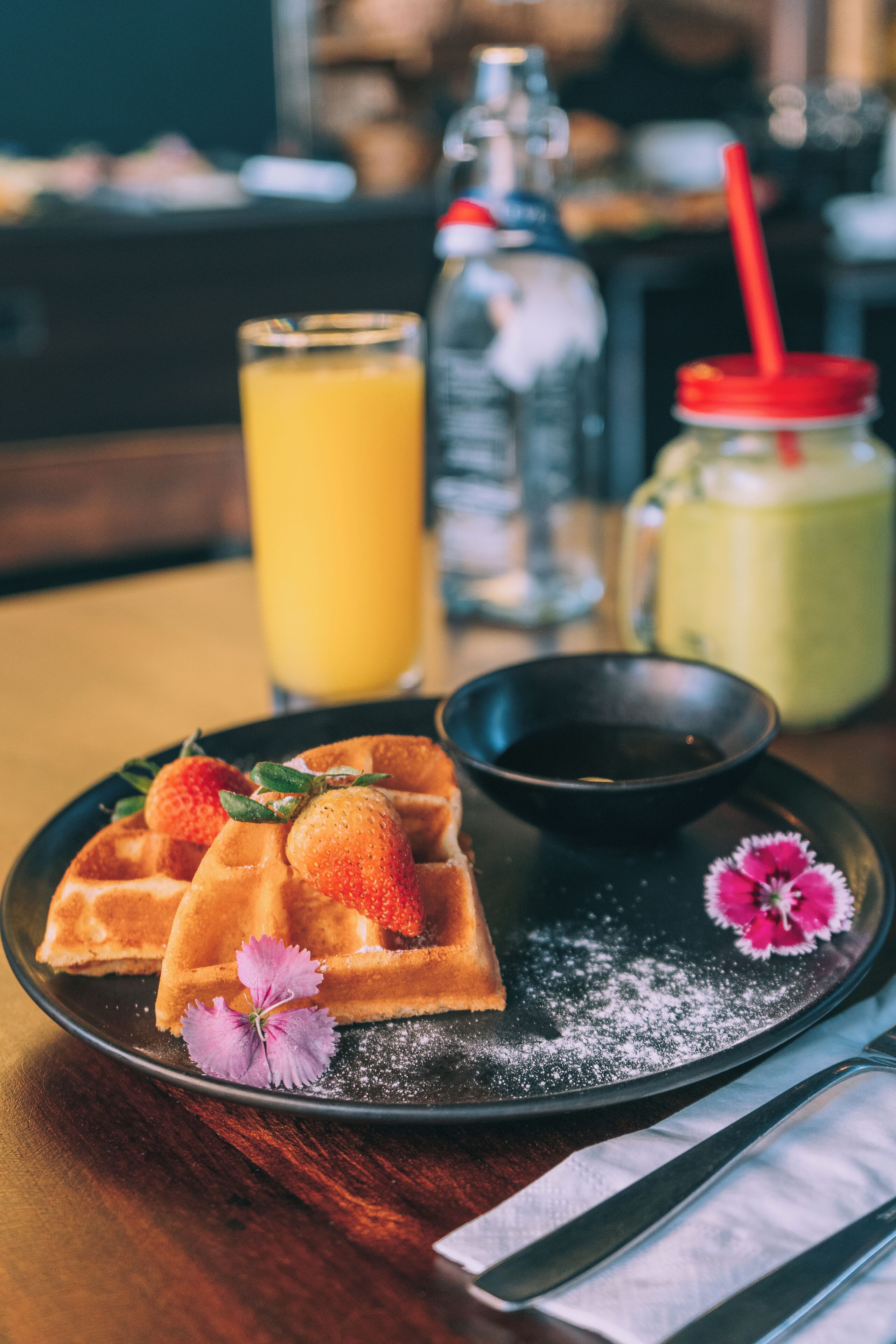 A plate of waffles with strawberries and a small bowl of syrup, a glass of orange juice, and a jar of jam on a wooden table.