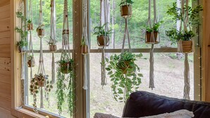 Une pièce dotée d’une grande fenêtre ornée de supports et de pots à plantes en macramé, qui offre une vue sur une zone boisée.
