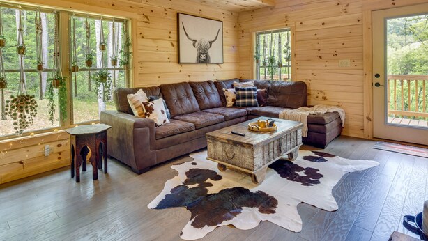 Un intérieur de chalet chaleureux avec un canapé d’angle en cuir marron, une table basse en bois et une grande fenêtre agrémentée de plantes.