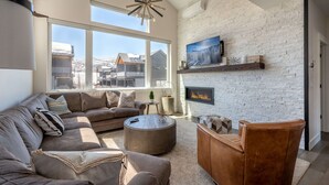Ein modernes Wohnzimmer mit einer Eckcouch, einem runden Couchtisch und einem Ledersessel. Ein Kamin ist in die weiße Ziegelwand integriert, und ein großes Fenster bietet einen Blick ins Freie.