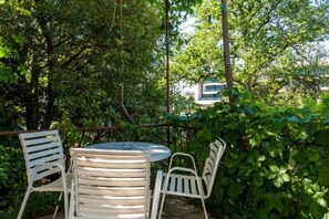 Ein runder Tisch mit vier weißen Stühlen in einem Garten, umgeben von dichtem Grün, mit einem Gebäude im Hintergrund.