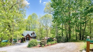 A rustic cabin with a gravel driveway, surrounded by dense trees and greenery. - Fast Wifi, covered porch, and fenced-in yard. Quiet neighborhood. Pet friendly! (Maggie Valley)