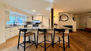 A modern kitchen with white cabinetry, a central island with bar stools, and a fireplace. - Luxury Redo: Hot Tub, Playroom, Fire-pit, Sleeps 12 (Egremont)