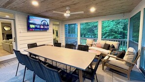Une pièce spacieuse avec une grande table et des chaises, un ensemble de canapés, un téléviseur fixé au mur et de grandes fenêtres donnant sur l’extérieur.