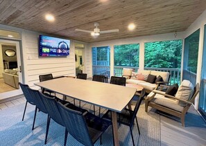 A spacious room with a large table and chairs, a sofa set, a TV mounted on the wall, and large windows offering a view of the outdoors.