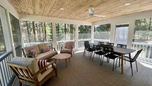 Un espace extérieur abrité, doté d’un plafond en bois, d’un ensemble de canapés, d’une table à manger avec des chaises et de grandes fenêtres offrant une vue sur les arbres.