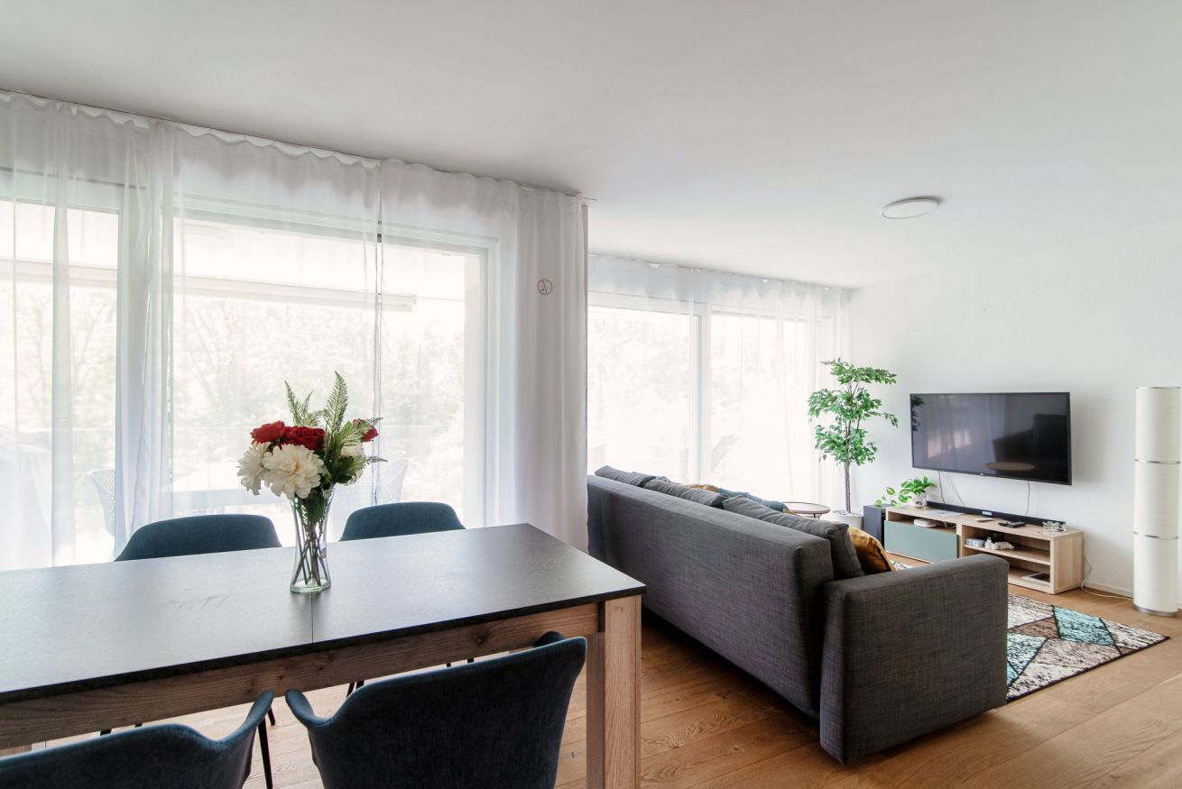 Ett modernt vardagsrum med ett matbord, en grå soffa, en tv och ett stort fönster med tunna, genomskinliga gardiner.