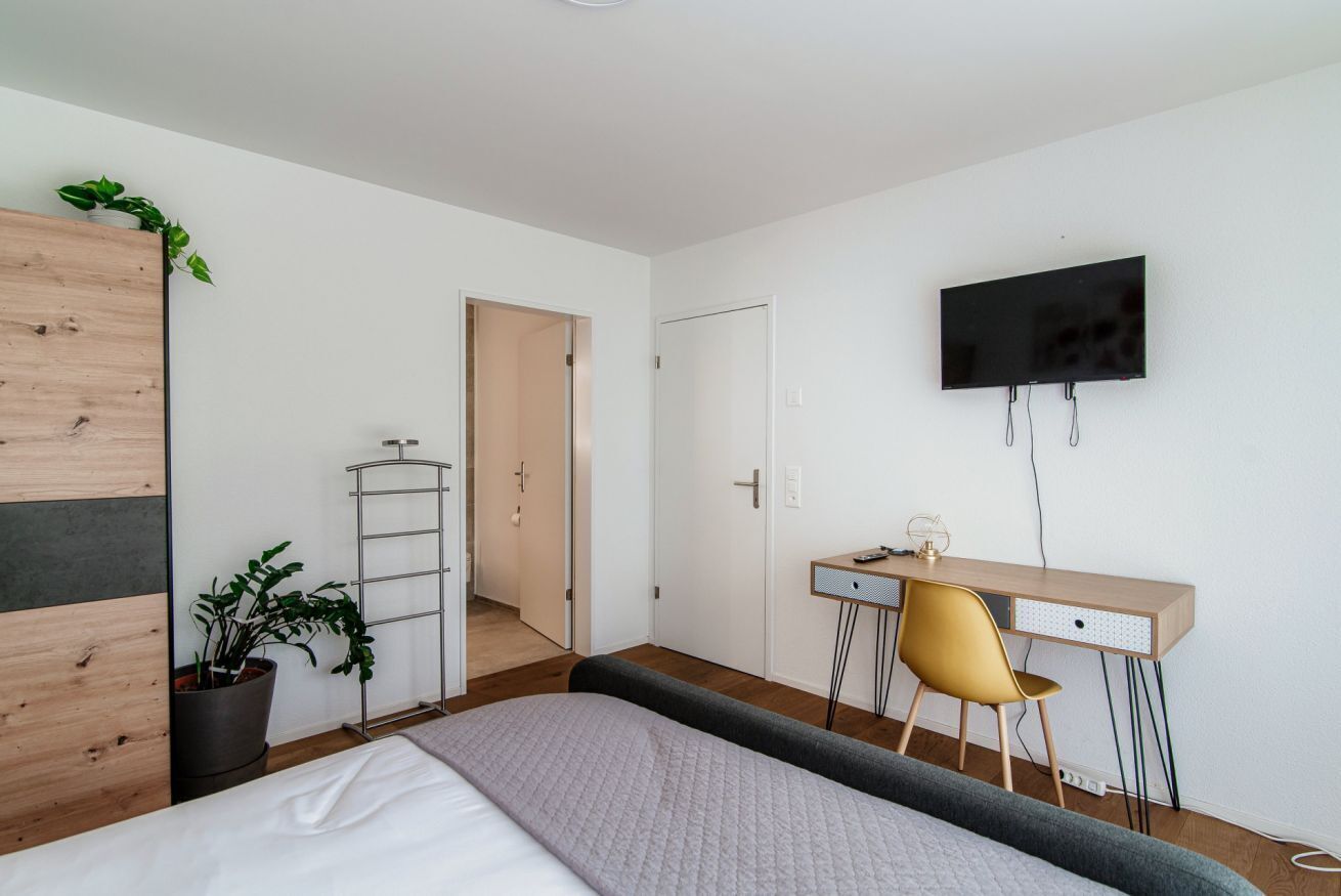 A bedroom with a bed, a desk, a chair, a TV, and a potted plant.