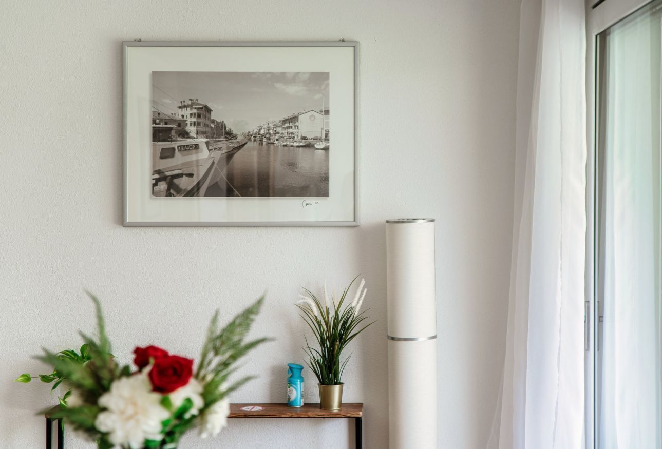 A framed black and white photograph of a canal with buildings, a small wooden shelf with a vase of flowers, and a tall white cylindrical object.