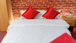 A hotel room with a brick wall, a bed with white bedding and red pillows, a wooden headboard, a nightstand with a water bottle, and a painting of flowers on the wall. - KAJI CHEN (Sunakothi)