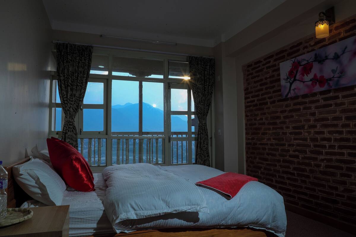A hotel room with a large window, a bed with white bedding, a red pillow, and a brick wall with a painting.