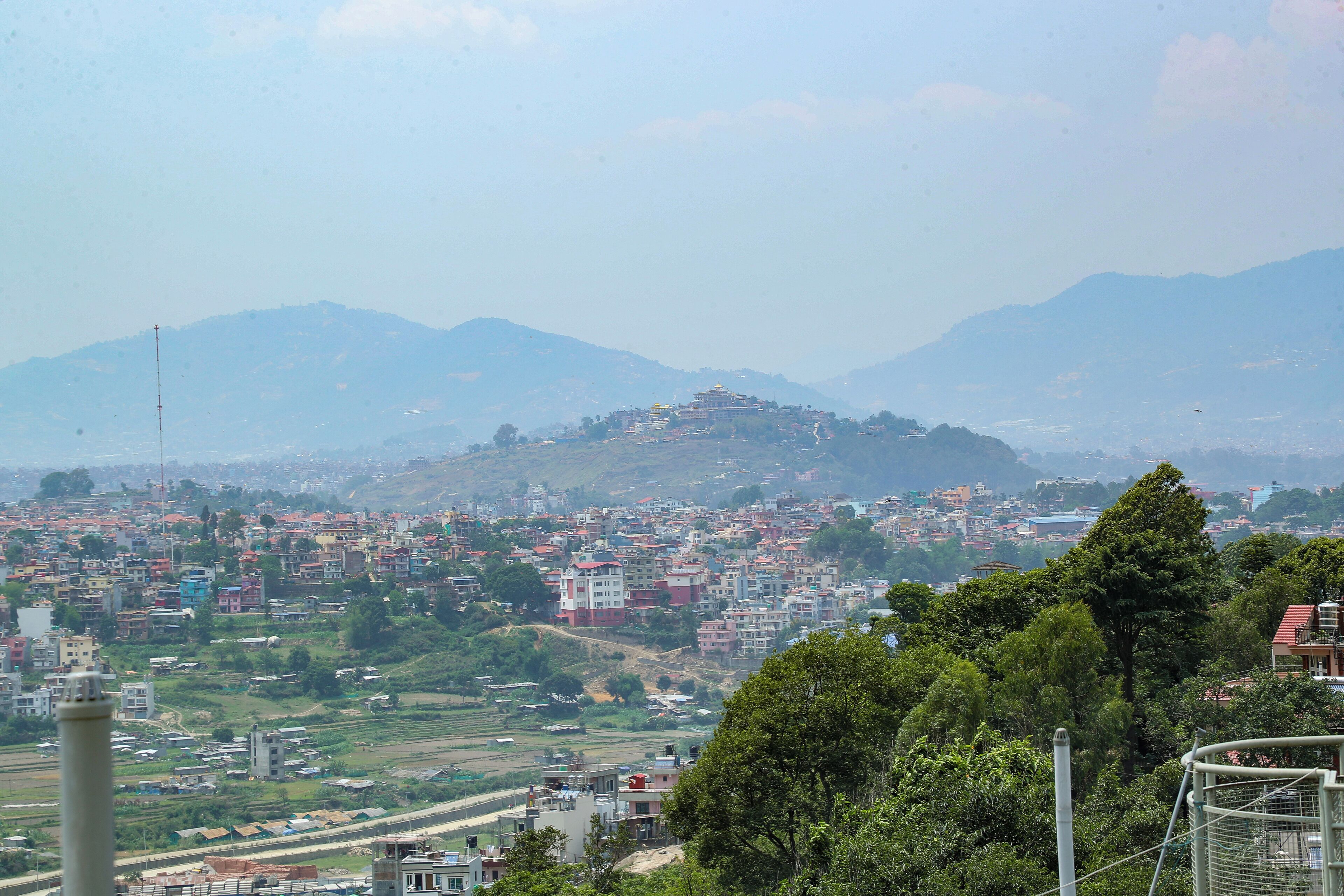 A panoramic view of a city with a prominent hilltop structure, possibly a temple or monument, surrounded by residential buildings and greenery.
