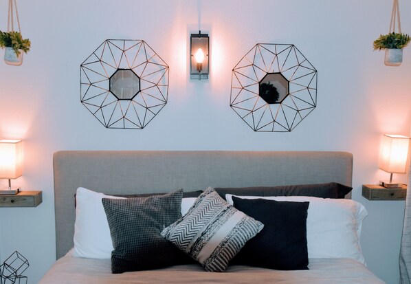 Ein Schlafzimmer mit einem Bett, Wandkunst mit geometrischen Formen, Nachttischlampen und einem Nachttisch mit einer Pflanze.