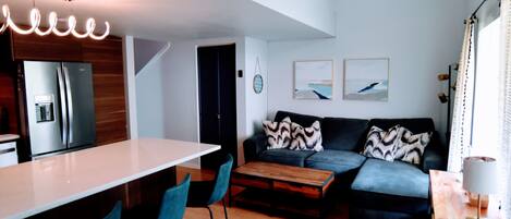 A modern kitchen with a refrigerator, a dining area with a table and chairs, a sofa with patterned cushions, and a coffee table.