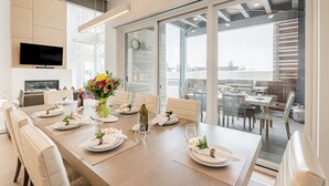Uma sala de jantar moderna com uma mesa comprida posta para seis pessoas, uma televisão montada na parede e vista da cidade através de grandes janelas.