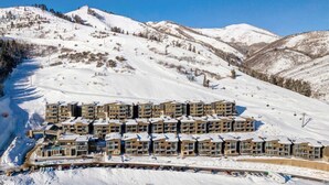 Estância de esqui coberta de neve com vários edifícios altos, provavelmente um hotel, apresentando uma fileira de quartos com varandas, ao fundo montanhas e pistas de esqui.