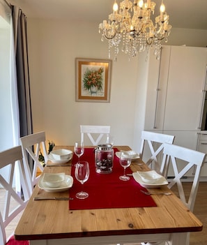Mesa de jantar posta para quatro pessoas, com cadeiras brancas, um jogo americano vermelho e um candelabro acima.