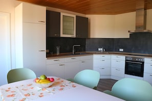 A compact kitchen with white cabinets, a black countertop, and a built-in oven. There is a table with a fruit bowl and two chairs. - Vacation apartment Hof Margarethental - Renovated half of the house for 6 persons on a beautiful (Goldelund)