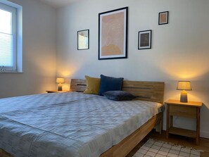 A bedroom with a large bed, two bedside tables with lamps, a window with blinds, and three framed pictures on the wall. - Haus Möwe - Ferienwohnung 5 "heringsmöwe" - Lietzow Appartementhaus Möwe (Lietzow)