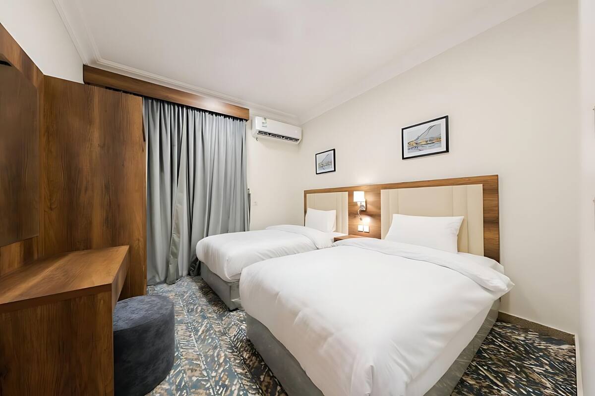 Quarto de hotel com duas camas, um guarda-roupa de madeira, uma escrivaninha e um tapete estampado.