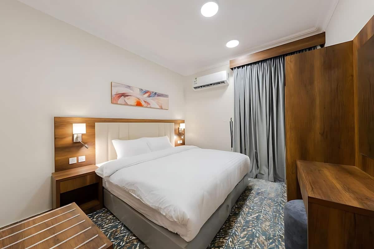 Quarto de hotel com uma cama grande, cabeceira de madeira, abajures, um quadro na parede e um banco de madeira.